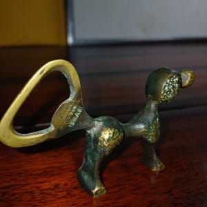 May include: A brass bottle opener shaped like a stylised four-legged animal with a dark, weathered patina and textured finish. The handle features an integrated loop and is stamped with the text PAL-BELL 714 ISRAEL on the side.