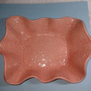 May include: A rectangular ceramic bowl with a glossy pink finish and a wavy, scalloped rim. The interior surface features a repeating embossed pattern of small, five-petaled flowers distributed evenly across the base and the sides of the dish.