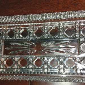 Peut inclure: Un plateau rectangulaire en verre transparent avec un motif pressé décoratif. Le bord présente une bordure crantée répétitive, tandis que la section intérieure affiche une série de formes géométriques octogonales autour d'un motif central en étoile.