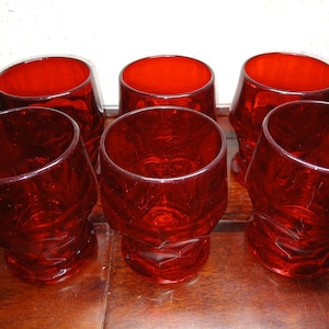 May include: A set of six matching red glass tumblers featuring a faceted, geometric pattern on the exterior. Each glass has a wide, circular rim and a sturdy base, showcasing a deep, translucent ruby red color throughout the entire glass structure.