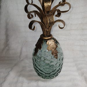 Puede incluir: Un objeto decorativo con forma de piña que presenta una base de vidrio transparente texturizado con un patrón de hoyuelos. La parte superior consta de hojas y un tallo de metal dorado, con detalles de hojas doradas adicionales en el vidrio.