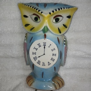 May include: A ceramic owl-shaped wall clock featuring a light blue body with yellow accents around the eyes and brown feather details. The clock face is white with black Roman numerals and two black hands, suspended by two thin metal chains.