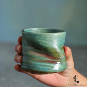 Könnte beinhalten: Ein kleiner, zylindrischer Keramikbecher mit einer glänzenden, mehrfarbigen Glasur in Blaugrün, Grün und Braun. Die Oberfläche weist eine subtile, gewellte Textur mit horizontalen Vertiefungen auf. Ein kreisförmiges Logo ist deutlich zu sehen.