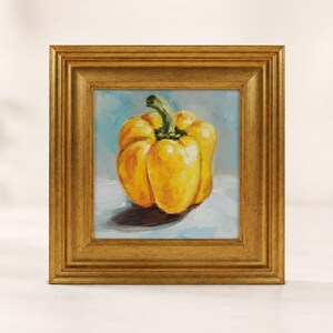 May include: An oil painting of a single yellow bell pepper with a green stem, rendered with visible brushstrokes. The pepper sits on a light surface against a soft blue background, all enclosed within a decorative, ornate gold-toned frame.