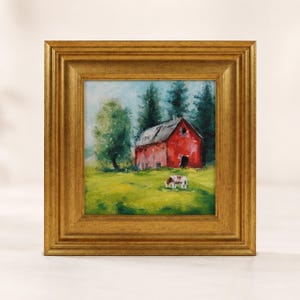 May include: An oil painting in a square gold frame depicting a vibrant red barn situated in a lush green field. The scene includes dark green pine trees in the background and two small horses grazing in the foreground under a light blue sky.