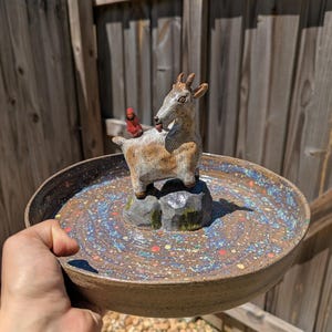 May include: A round ceramic bowl with a speckled brown, blue, and multi-coloured glaze. Centered inside the bowl is a sculpted clay goat standing on two grey rocks, with a small red cardinal bird perched on the goat's back, facing away.