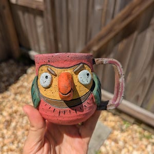 May include: A handmade ceramic mug shaped like a parrot with a speckled pink body, a yellow face, a prominent orange beak, and green wing accents. The mug features a wide, flat handle and a textured surface with a cartoonish, expressive face.