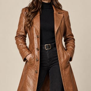 May include: A tan leather coat featuring a notched lapel, long sleeves, and a four-button front closure. The coat has two side pockets and a structured fit. It is worn over a black ribbed turtleneck sweater and black jeans with a black belt.