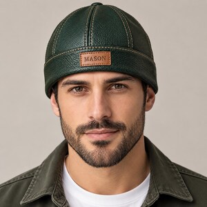 Puede incluir: Un gorro tipo docker de cuero texturizado en color verde oscuro con una corona redondeada, costuras verticales y borde enrollado. Un parche rectangular de cuero color canela está centrado en el borde frontal con la palabra MASON en relieve.