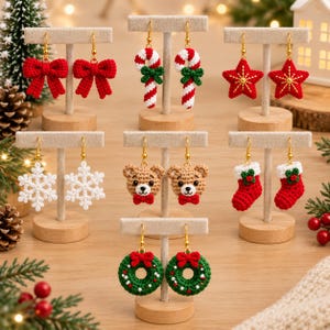 Peut inclure: Sept paires de boucles d'oreilles de Noël en crochet faites à la main sur des supports en T en bois. Les motifs incluent des nœuds rouges, des sucres d'orge, des étoiles, des flocons blancs, des oursons, des chaussettes et des couronnes.