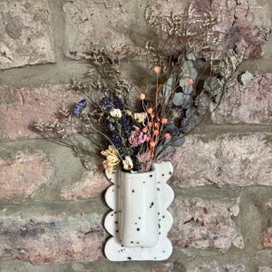 Op de afbeelding: Een witte keramische wandvaas met een gespikkeld patroon en een geschulpte rand, gevuld met een boeket gedroogde bloemen waaronder eucalyptus, lavendel en kleine oranje bessen, gemonteerd tegen een muur van gestructureerde rode baksteen.