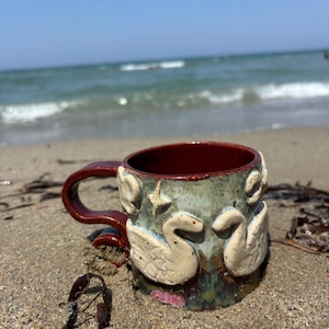 May include: A ceramic mug with a dark red interior and handle, featuring a light green exterior decorated with raised white swan figures and small star shapes. The mug has a textured surface and is partially embedded in the sandy beach ground.