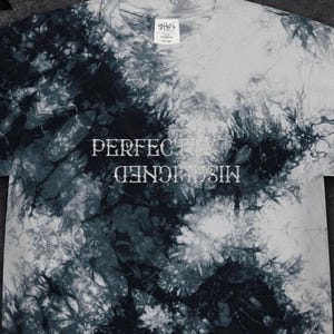 May include: A short-sleeve t-shirt featuring a black and white tie-dye pattern. The center of the chest displays the text "PERFECTLY" in white, with the word "MISALIGNED" printed directly below it in an inverted, upside-down orientation.