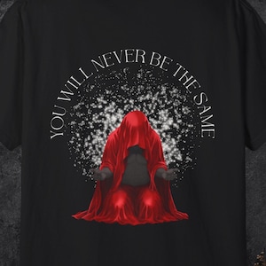 May include: A black t-shirt featuring a graphic of a figure draped in a vibrant red hooded cloak, kneeling with hands outstretched. Behind the figure is a white speckled pattern. The text "YOU WILL NEVER BE THE SAME" arches above the figure.