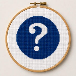 以下が含まれることがあります： 木製の円形刺繍枠に白い布が張られ、クロスステッチのデザインが施されています。デザインは、大きな無地の濃い青色の円の中に、個々のステッチされた正方形で構成された白い疑問符が中央に配置されています。