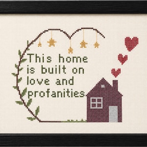 Peut inclure: Une broderie au point de croix encadrée sur tissu crème avec le texte This home is built on love and profanities en vert foncé. Le motif comprend une maison violet foncé avec des cœurs rouges sortant de la cheminée et des étoiles dorées.
