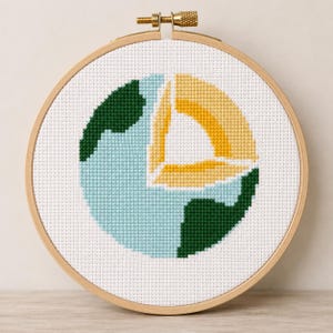 May include: A circular wooden embroidery hoop containing a cross-stitch pattern of the Earth. The design features light blue oceans, dark green landmasses, and a yellow wedge-shaped cutout revealing the planet's internal layers on a white fabric.