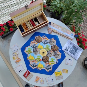 Pode incluir: Um conjunto de jogo de tabuleiro Settlers of Catan com um tabuleiro hexagonal de peças de terreno, uma caixa de madeira com peças coloridas, um suporte para cartas, um manual de regras e várias cartas sobre uma mesa redonda branca.