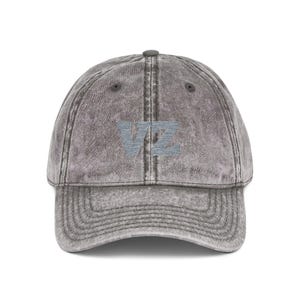 Puede incluir: Una gorra de béisbol gris carbón con un acabado desgastado y lavado al ácido. El panel frontal presenta las letras VZ bordadas en hilo gris claro. La gorra tiene una visera curva, un botón en la parte superior y ojales cosidos.