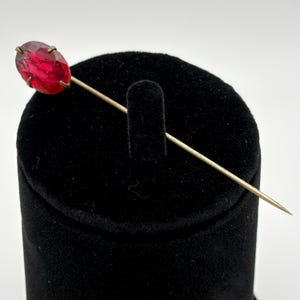 May include: A stick pin featuring a faceted, oval-shaped red gemstone set in a metallic prong setting. The long, slender metal pin extends from the base of the stone, showcasing a polished finish and a sharp, pointed end for securing into fabric.