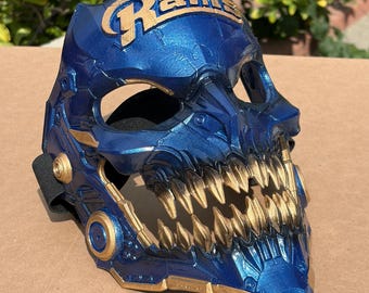 May include: A metallic blue and gold skull-shaped mask featuring the word Rams in gold lettering across the forehead. The mask has sharp, pointed gold teeth, circular side accents, and a black elastic strap attached to the sides for securing it.