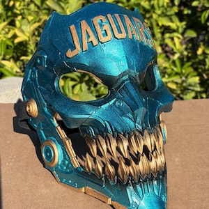 May include: A teal and gold mask featuring a mechanical, skull-like design with sharp, pointed teeth. The word JAGUARS is embossed in gold lettering across the forehead area. The mask includes circular gold accents on the sides and a black strap.