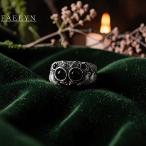 May include: A silver ring shaped like a jumping spider with textured fur-like detailing and four prominent black circular eyes. The word FAELYN is printed in white text in the upper left corner of the frame, above the spider-shaped ring.