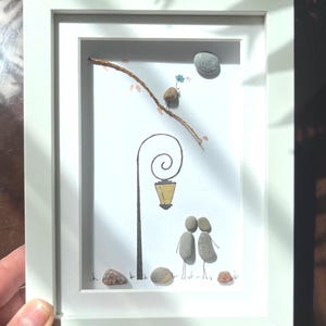 Op de afbeelding: Een ingelijst kunstwerk van kiezelstenen met twee figuren onder een getekende lantaarnpaal. Een kleine vogel van een kiezelsteen zit op een tak en houdt een blauwe bloem vast. Een grijze kiezelsteenmaan in de hoek, met kleine steentjes op de grond.