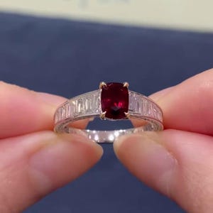 Könnte beinhalten: Ein Ring mit einem zentralen, kissenförmigen, tiefroten Rubin, der in gelbgoldenen Krappen gefasst ist. Die Ringschiene besteht aus poliertem weißem Metall und ist mit klaren, rechteckigen Baguette-Diamanten im Kanalbesatz versehen.