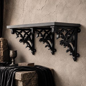 May include: A black wall-mounted shelf supported by three ornate, intricate brackets featuring carved gargoyle-like figures and openwork scroll designs. The shelf has a flat, rectangular top surface and is mounted horizontally against a textured wall.