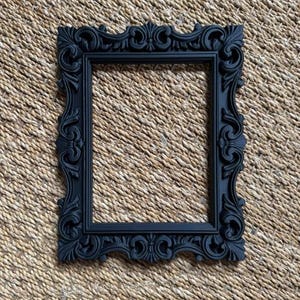 May include: A rectangular picture frame in a matte black finish, featuring an ornate, raised relief pattern along the entire border. The frame has a hollow center and a symmetrical design with scrolling leaf-like motifs and decorative flourishes.