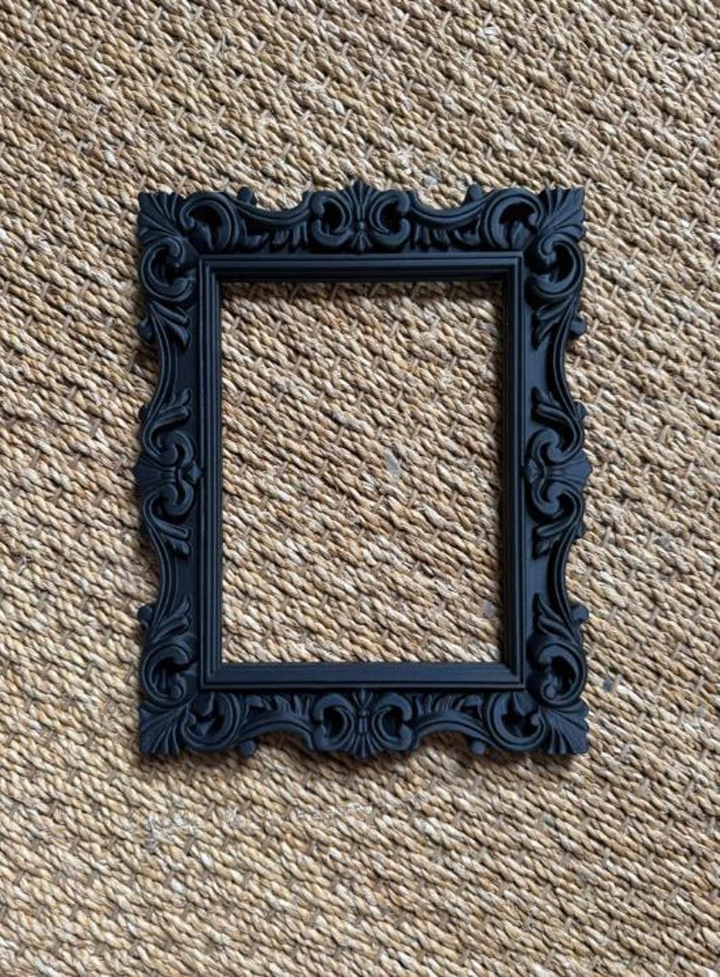 May include: A rectangular picture frame in a matte black finish, featuring an ornate, raised relief pattern along the entire border. The frame has a hollow center and a symmetrical design with scrolling leaf-like motifs and decorative flourishes.
