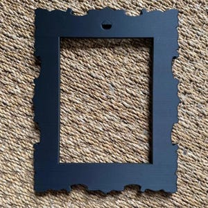 May include: A rectangular picture frame with a matte black finish and ornate, scalloped edges. The frame features a central rectangular opening and a small, triangular mounting hole at the top center for hanging on a wall or display surface.