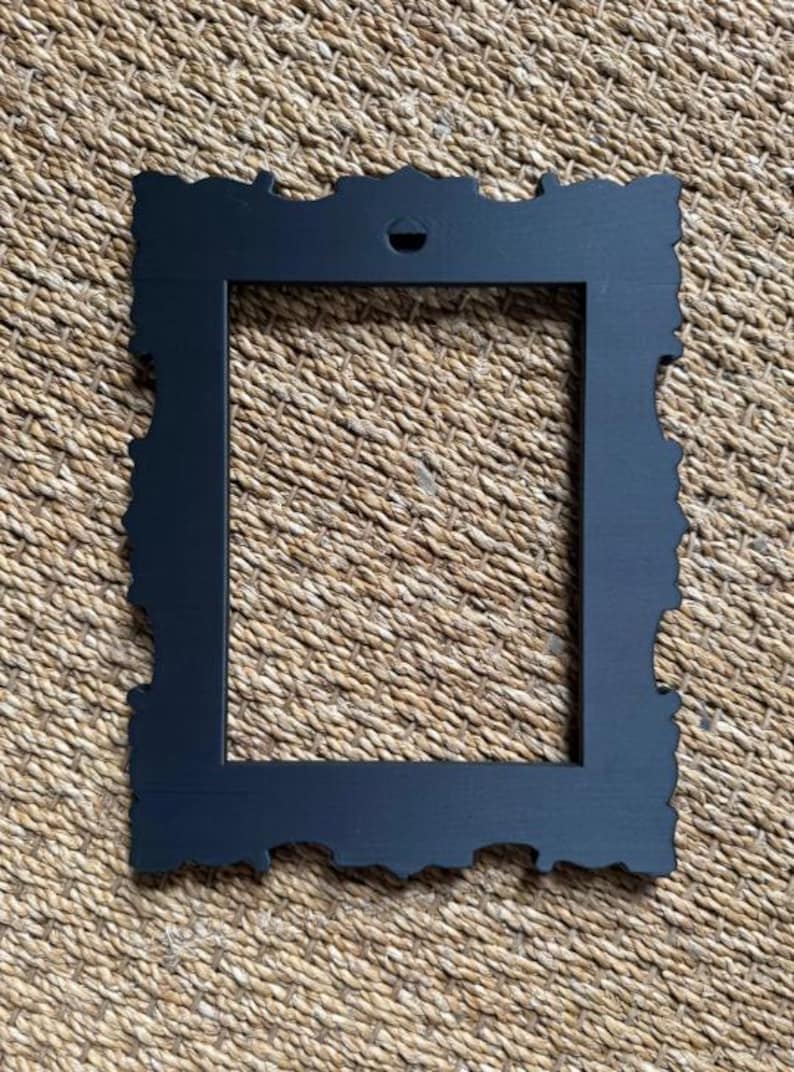 May include: A rectangular picture frame with a matte black finish and ornate, scalloped edges. The frame features a central rectangular opening and a small, triangular mounting hole at the top center for hanging on a wall or display surface.