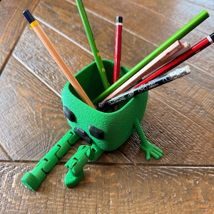 Peut inclure: Un porte-crayons vert vif imprimé en 3D en forme de personnage cubique avec des jambes et des bras articulés. Le récipient contient plusieurs crayons assortis, notamment des verts, rouges et noirs, avec diverses gommes et inscriptions.