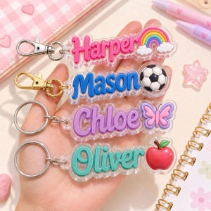 May include: Four acrylic keychains with raised lettering and colorful icons. The first reads Harper with a rainbow, the second Mason with a soccer ball, the third Chloe with a butterfly, and the fourth Oliver with a red apple. Each has a metal clasp.