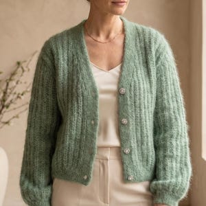 Puede incluir: Un cárdigan de punto acanalado en color verde salvia, de manga larga, con escote en V y cierre frontal de cuatro botones redondos decorativos. La prenda se lleva sobre una camiseta de tirantes y pantalones de color crema.