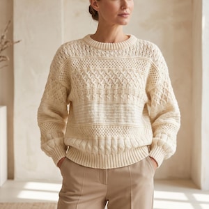Könnte beinhalten: Ein cremefarbener Langarmpullover mit Rundhalsausschnitt und einem komplexen Strickmuster. Das Design umfasst horizontale Bänder aus Zopfstrick, Wabenmuster und gerippten Texturen, die sich über den Brustbereich und die Ärmel erstrecken.