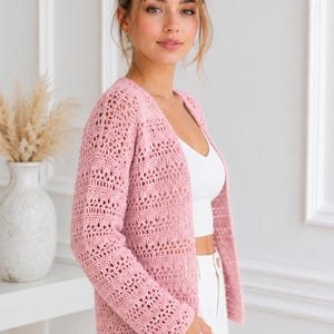 May include: A light pink open-front cardigan featuring a textured crochet knit pattern with long sleeves and a straight hemline. The garment is worn over a plain white crop top and white trousers, showcasing a detailed, repeating openwork stitch design.