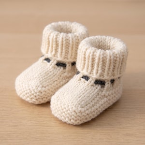Könnte beinhalten: Ein Paar cremefarbene, gestrickte Babyschuhe mit einem dicken Rippbündchen und einer dezenten, dunkelgrauen horizontalen Akzentlinie, die direkt unter dem Knöchel eingearbeitet ist. Die Schuhe haben eine grobe Strickstruktur und eine runde Form.