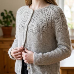 Könnte beinhalten: Eine hellgraue Langarm-Strickjacke mit Rundhalsausschnitt und durchgehender Knopfleiste. Das Kleidungsstück ist im Schulterbereich mit einem strukturierten Strickmuster versehen, das filigrane, spitzenartige Details auf der Brust aufweist.