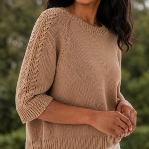 Könnte beinhalten: Ein beigefarbener Strickpullover mit Rundhalsausschnitt, Dreiviertelärmeln und geripptem Saum. Die Ärmel sind mit einem vertikalen Zopfstrickmuster entlang der Außenseite versehen, während der Körper eine glatte, schlichte Strickstruktur aufweist.