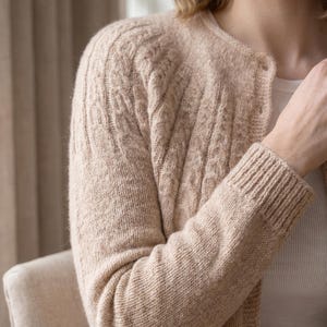Könnte beinhalten: Eine beigefarbene Strickjacke mit Rundhalsausschnitt, langen Ärmeln mit gerippten Bündchen und einer Knopfleiste auf der Vorderseite. Das Kleidungsstück zeigt ein strukturiertes Zopfmuster an den Schultern und den oberen Ärmeln.