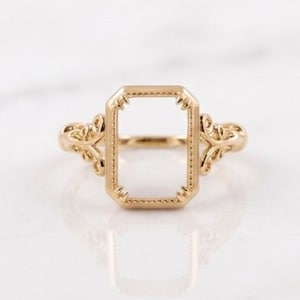 May include: A gold ring featuring a central rectangular frame with a beaded edge and decorative filigree detailing on the sides of the band. The center of the rectangular frame is open, showcasing a hollow design with intricate metalwork.