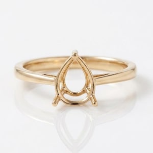 May include: A gold ring setting featuring a pear-shaped prong mount designed to hold a single gemstone. The band is smooth and polished, showcasing a classic metal construction with a central basket structure intended for a teardrop-shaped stone.