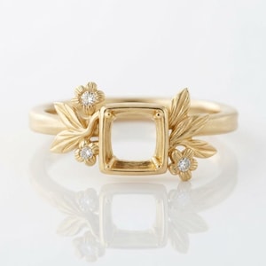 May include: A gold ring featuring a central square setting for a gemstone, flanked by sculpted gold leaves and three small, round, clear inset diamonds. The metal has a matte finish, and the design incorporates floral motifs on both sides.
