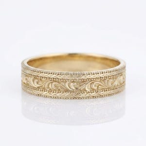 Op de afbeelding: Een gouden ring met een centrale band van ingewikkelde krulgravures, geflankeerd door twee dunnere randen met gestructureerde, herhalende bladachtige patronen. Het metaal heeft een gepolijste afwerking met gedetailleerd reliëfwerk over het oppervlak.
