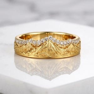 May include: A gold ring featuring a textured mountain range relief design around the band. The upper edge of the ring is accented with a row of small, clear, round-cut stones that follow the jagged peaks of the mountain landscape motif.