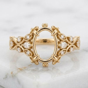 May include: A gold ring featuring an empty oval center setting surrounded by a decorative filigree frame. The band is adorned with small, clear, round and pear-shaped stones embedded along the intricate, symmetrical metalwork design.