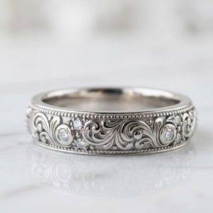 Könnte beinhalten: Ein silberfarbener Metallring mit einem komplizierten, gravierten Rankenmuster auf dem Band. Das Design enthält kleine, runde, klare facettierte Steine, die in das geschwungene Relief eingebettet sind und von einem feinen Perlenrand umgeben sind.